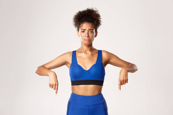 Concept van training en fitness. Droevig en somber Afrikaans-Amerikaans meisje athelte, het dragen van blauwe sportkleding, wijzende vingers naar beneden en snikkend, witte achtergrond - Foto, afbeelding