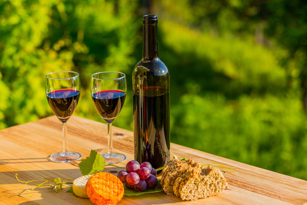 Weinflasche, Trauben, Käse und Brot, auf Holztisch, im Freien - Foto, Bild