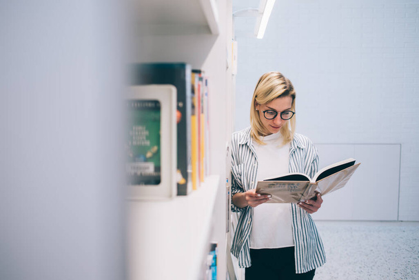 Young woman in optical eyeglasses and smart casual wear holding literature interested on plot near bookcase, beautiful Caucasian hipster girl reading novel book from library collection on shelves - Photo, Image