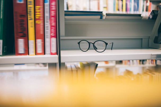 Selective focus on trendy optical spectacles for provide eyes protection staying on shelf bookcase in university library, stylish classic eyewear for vision correction near literature best sellers - Fotoğraf, Görsel