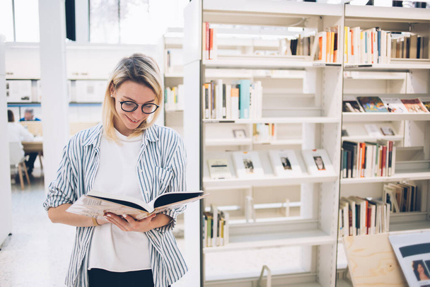 Cheerful young woman in optical eyewear reading information from knowledge textbook preparing for seminar in library, pretty student dressed in casual wear holding book in hands and smiling - Foto, Bild