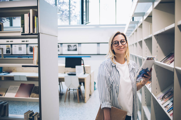 Half length portrait of cheerful youthful woman in optical eyeglasses for vision correction smiling at camera while selecting literature book from bookcase in public library, education concept - Foto, Imagem