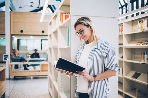 Side view of young woman with blonde hair holding book in hands and reading interesting information in university library, happy female in glasses learning literature plot to improve knowledge skills - Foto, imagen