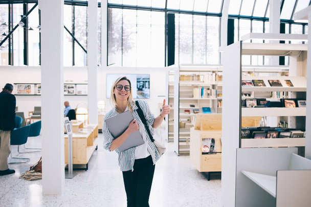 Μισού μήκους πορτρέτο της ευτυχούς φοιτήτριας με οπτικά γυαλιά για την προστασία των ματιών χαμογελώντας στην κάμερα και δείχνοντας τον αντίχειρα επάνω στο πανεπιστήμιο Εθνική βιβλιοθήκη, χαρούμενος Καυκάσιος ελεύθερος επαγγελματίας με φορητό υπολογιστή - Φωτογραφία, εικόνα