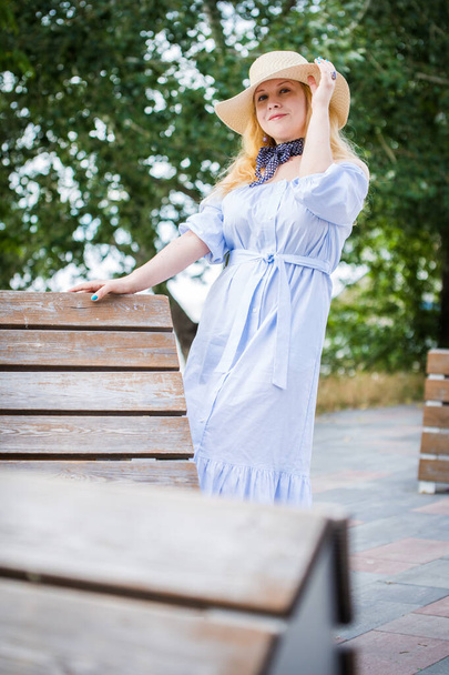 blonde in a blue dress and a beautiful hat posing on a bench in a city park - Φωτογραφία, εικόνα