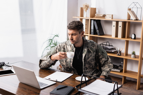 Militärangehöriger in Uniform hält Becher in der Hand, während er auf Laptop schaut  - Foto, Bild