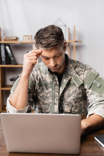 pensive soldier in uniform sitting at desk and using laptop  - 写真・画像