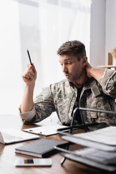 překvapený voják v uniformě drží pero v blízkosti notebooku s chytrým telefonem na rozmazané popředí - Fotografie, Obrázek