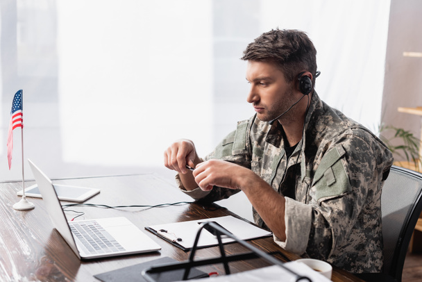 Πατριώτης στρατιωτικός με ακουστικά που κρατάει στυλό και κοιτάζει το λάπτοπ κοντά στην αμερικανική σημαία  - Φωτογραφία, εικόνα