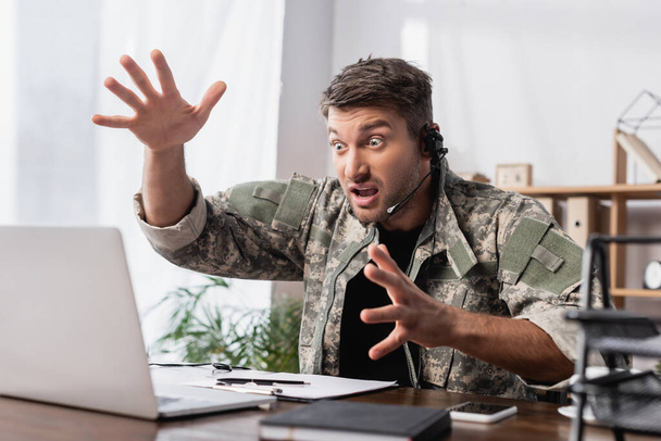 zszokowany wojskowy w mundurze i słuchawkach gestykulujący przy laptopie - Zdjęcie, obraz