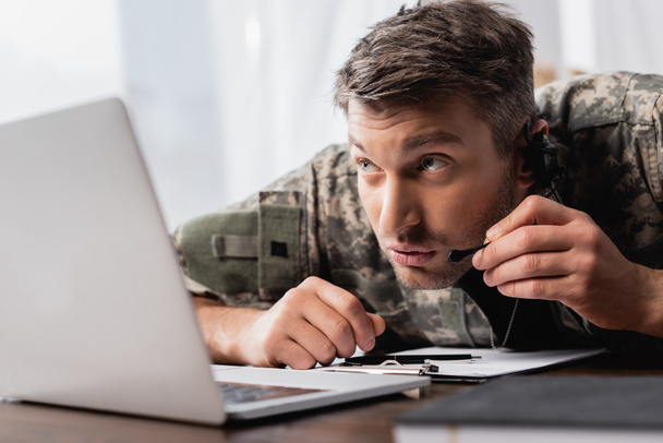 Militärmann in Uniform flüstert in Mikrofon auf Headset neben Laptop - Foto, Bild