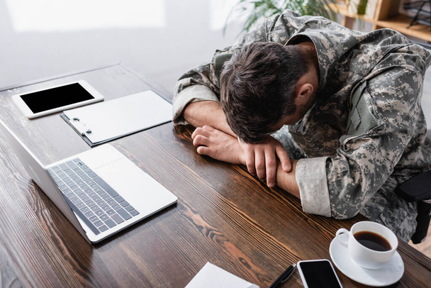 uomo militare in uniforme sdraiato sulla scrivania vicino al computer portatile e gadget con schermo bianco - Foto, immagini