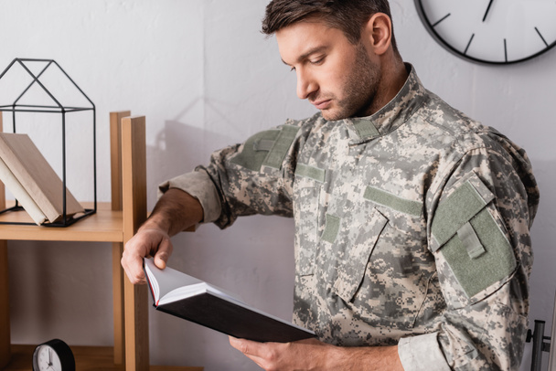 militar serio en uniforme sujetando bloc de notas cerca de estante de madera - Foto, imagen