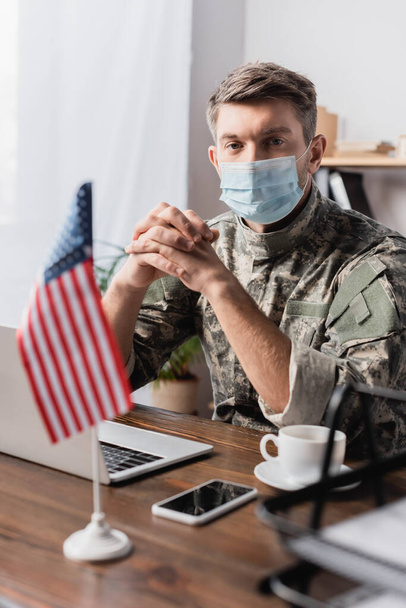 militar en uniforme y máscara médica sentado con las manos apretadas cerca de gadgets y bandera americana en primer plano borrosa  - Foto, imagen