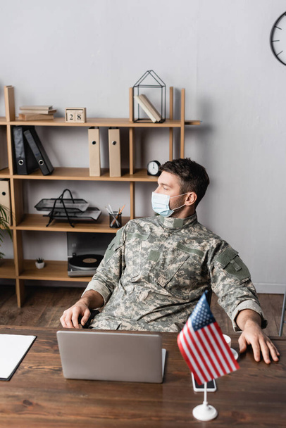 militar en uniforme y máscara médica sentado cerca de dispositivos y bandera americana en el escritorio - Foto, imagen