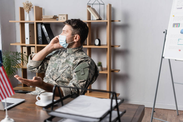 military man in uniform and medical mask talking on smartphone near american flag on desk - Foto, afbeelding