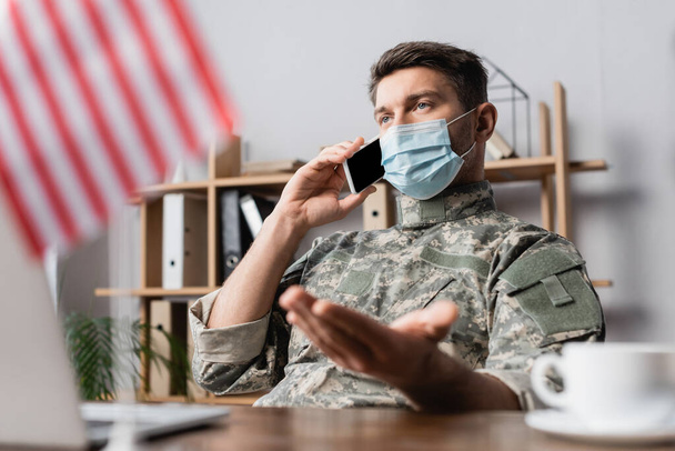 militar en máscara médica hablando en el teléfono inteligente cerca de bandera americana en primer plano borrosa - Foto, Imagen