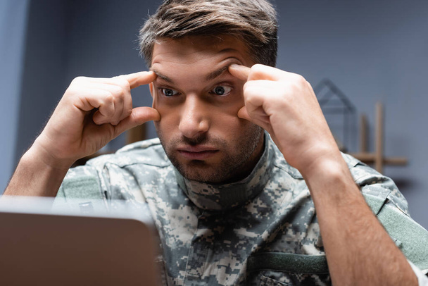 vermoeide militair in uniform kijkend naar laptop op wazige voorgrond - Foto, afbeelding