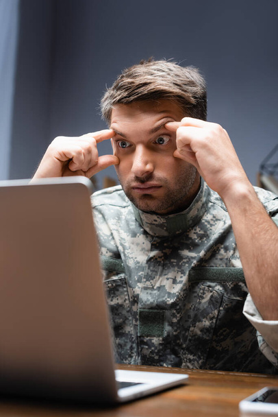 измученный военный в форме смотрит на ноутбук на размытом переднем плане - Фото, изображение