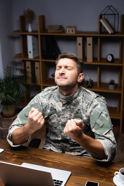 vzrušený voják v uniformě sedí se zaťatými pěstmi a raduje se z notebooku a smartphonu na stole - Fotografie, Obrázek