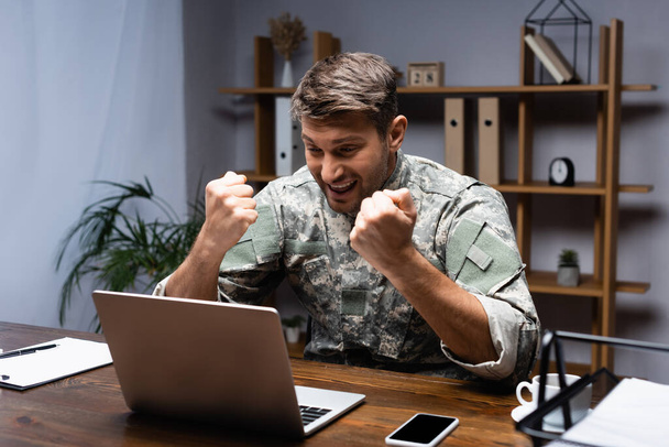 izgatott katona ül összeszorított ököllel, miközben örül közelében laptop és okostelefon az asztalon - Fotó, kép