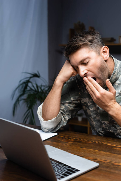 εξαντλημένος στρατιωτικός με στολή που καλύπτει το στόμα ενώ χασμουριέται κοντά στο λάπτοπ - Φωτογραφία, εικόνα