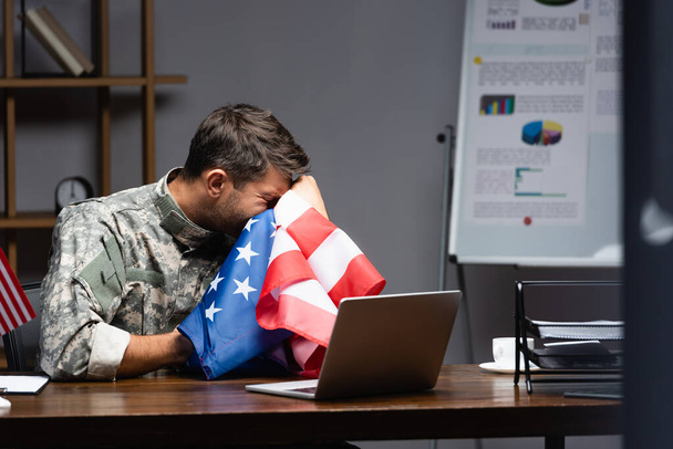 στρατιωτικός με στολή που κλαίει και καλύπτει το πρόσωπο με σημαία της Αμερικής κοντά στο laptop  - Φωτογραφία, εικόνα