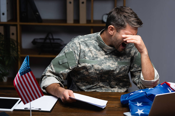 Aufgebrachter Soldat in Uniform, der Augen verdeckt und Brief hält, während er in der Nähe von Geräten und amerikanischer Flagge schreit  - Foto, Bild