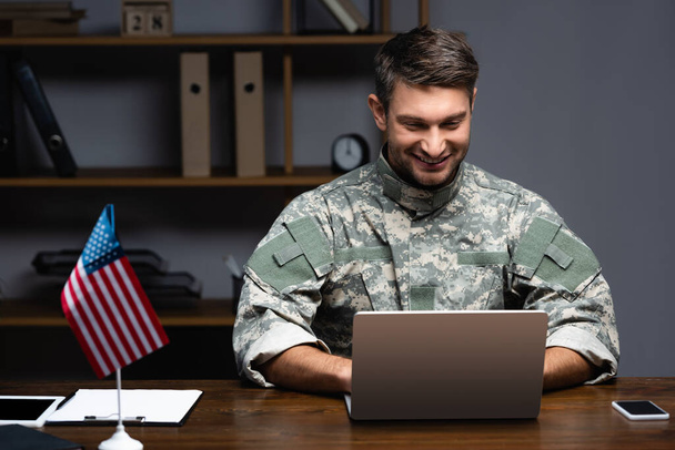 felice uomo militare in uniforme utilizzando laptop vicino bandiera americana e gadget  - Foto, immagini