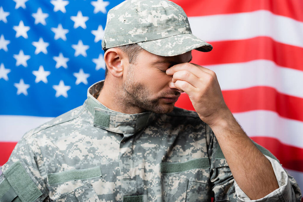 szomorú katona egyenruhás és sapkás törölgeti könnyeit, miközben sír közelében amerikai zászló elmosódott háttér - Fotó, kép