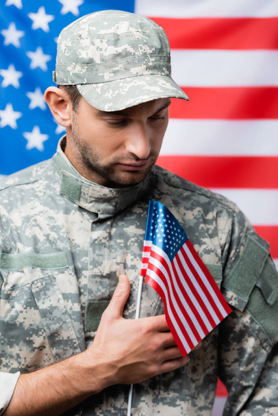 patriotic military man in uniform and cap holding small american flag on blurred background - Photo, Image