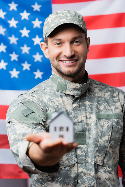 feliz militar en uniforme celebración casa modelo cerca de la bandera americana sobre fondo borroso - Foto, Imagen