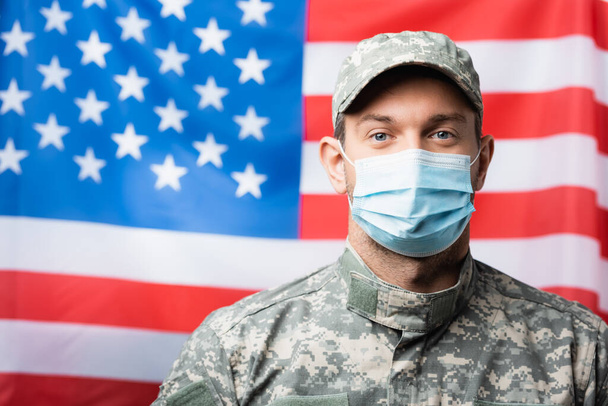 patriotique militaire en uniforme et masque médical près du drapeau américain sur fond flou - Photo, image