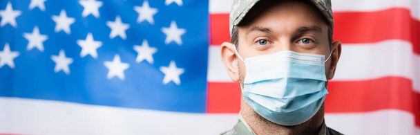 patriotický voják v uniformě a lékařské masce poblíž americké vlajky na rozmazaném pozadí, prapor - Fotografie, Obrázek