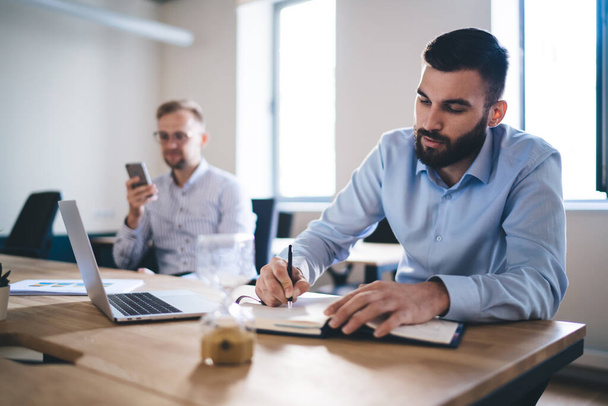Caucasian man in formal t shirt writing strategy ideas in textbook sitting at table desktop with male colleague using smartphone gadget for phoning on blurred background, office working process - Photo, Image