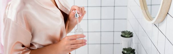 Vue recadrée de la femme en peignoir de soie tenant sérum cosmétique dans la salle de bain, bannière - Photo, image