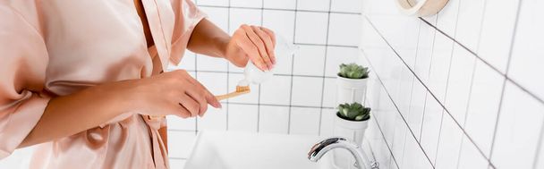 Vista recortada de la mujer en bata de satén aplicando pasta de dientes en el cepillo de dientes en el baño, pancarta  - Foto, Imagen