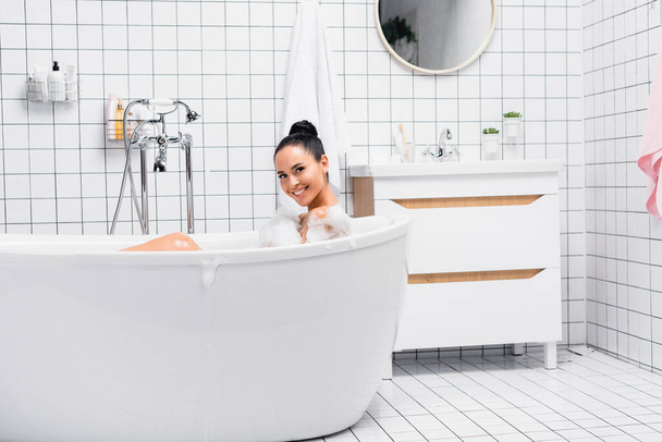 Cheerful brunette woman looking at camera while taking bath with foam  - Φωτογραφία, εικόνα