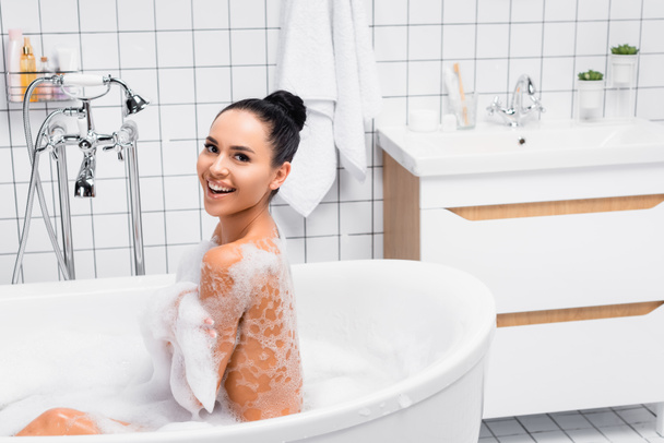 Brunette woman in foam smiling at camera while taking bath at home  - Foto, Imagem