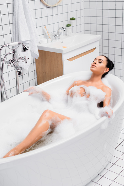 Sensual brunette woman lying with closed eyes while taking bath at home  - Foto, afbeelding