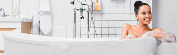 Привлекательная женщина улыбается, сидя в ванной с пеной в современной ванной комнате, баннер  - Фото, изображение