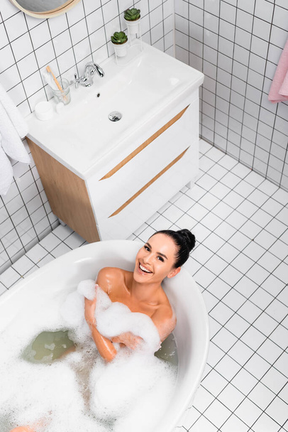 Horní pohled na veselou ženu usmívající se na kameru při ležení ve vaně s prádlem  - Fotografie, Obrázek