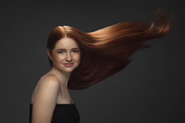 Beautiful model with long smooth, flying red hair isolated on dark studio background. - Foto, Imagem