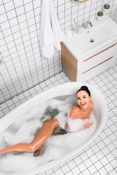 Vista superior de la mujer morena sonriendo a la cámara mientras toma un baño con espuma en el baño moderno  - Foto, Imagen