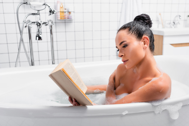 Чувственная женщина читает книгу, сидя в ванной с пеной в современной ванной комнате  - Фото, изображение