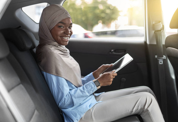 Cherful zwarte vrouw in hidjab met behulp van digitale tablet op de achterbank in de auto - Foto, afbeelding