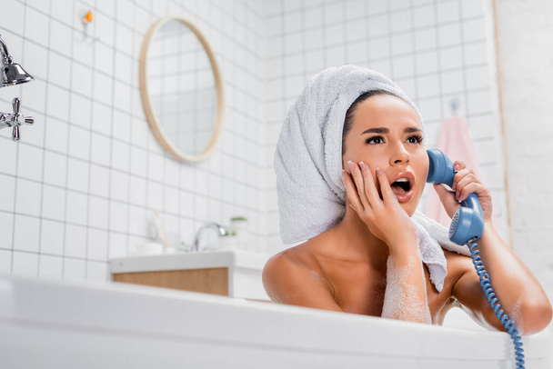 Shocked woman in towel talking on telephone while sitting in bathtub at home  - Foto, immagini