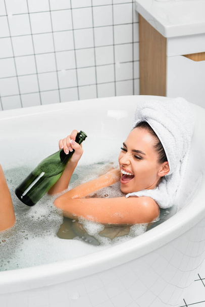 Excited woman with towel on head holding bottle of champagne in bath with foam  - Fotó, kép