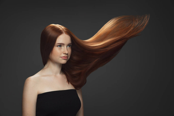 Beautiful model with long smooth, flying red hair isolated on dark studio background. - 写真・画像