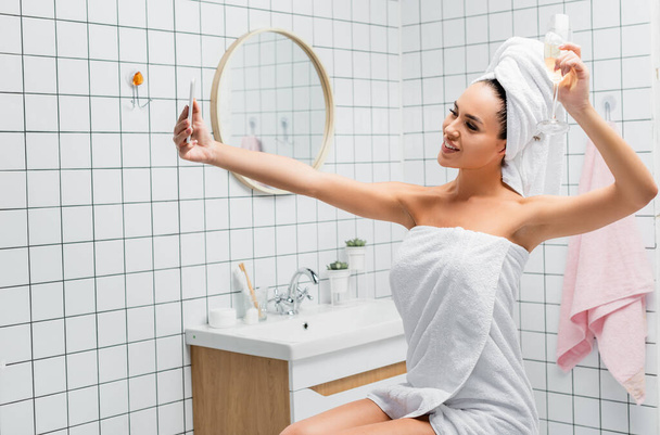 Usmívající se žena v ručnících drží sklenici šampaňského při užívání selfie v koupelně  - Fotografie, Obrázek